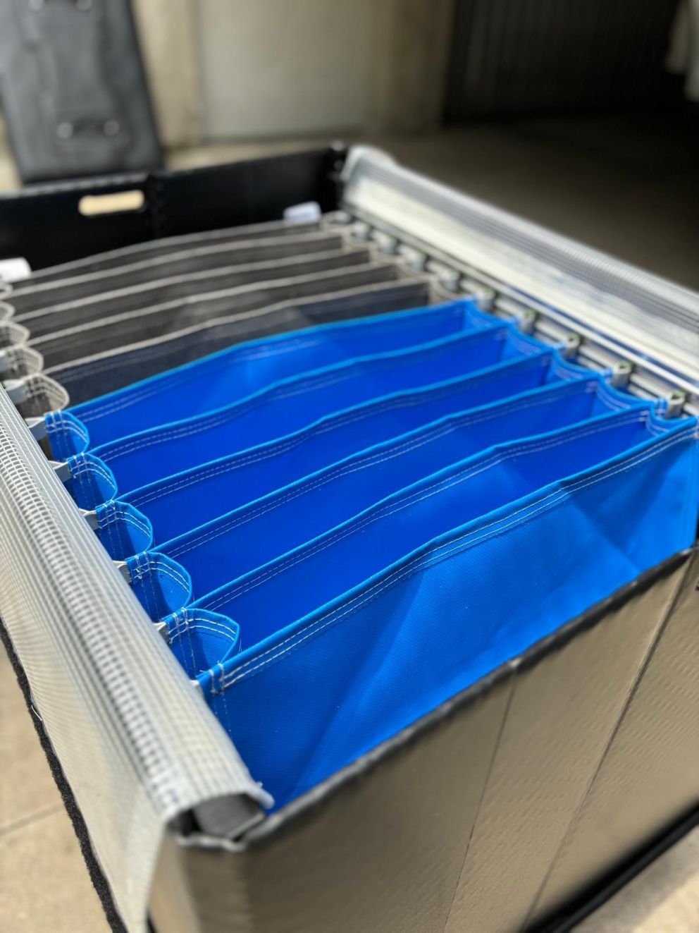 A close-up view of a multi-compartment storage container featuring several blue and black fabric sections, with a metal frame and a textured surface, set against a blurred background.