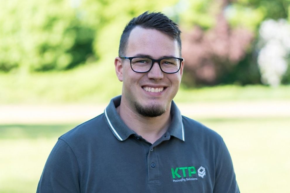 Smiling person wearing glasses and a gray polo shirt with a logo, standing outdoors against a blurred green background.