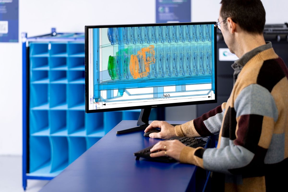 A person working at a desk looking at a monitor displaying a 3D design with colorful components, surrounded by blue storage units in a modern workspace.
