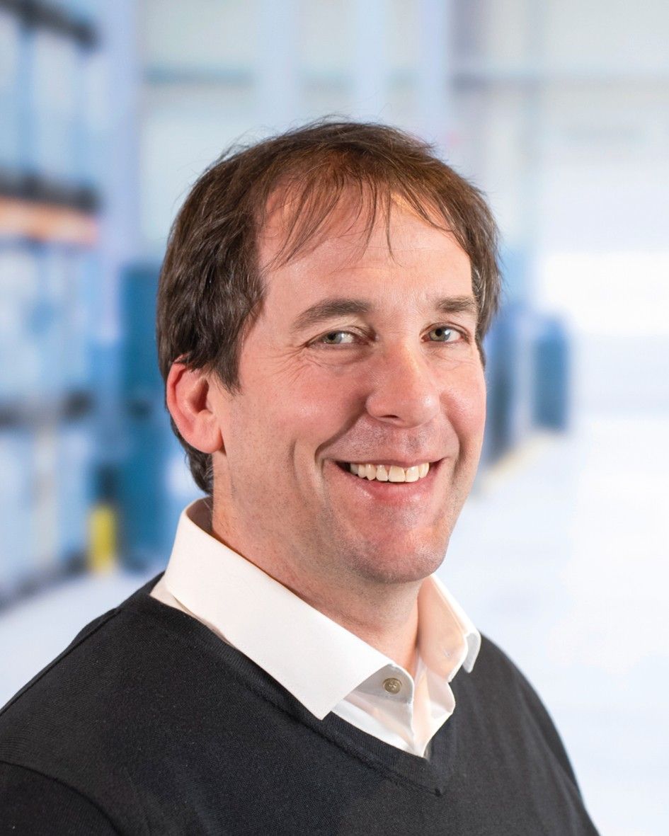 Smiling man wearing a black sweater over a white shirt, set against a blurred industrial background.