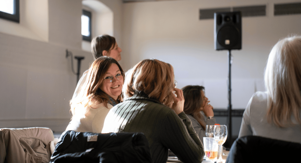 A group of people seated at a table in a bright, airy room, with a woman smiling in the foreground, and speakers visible in the background.