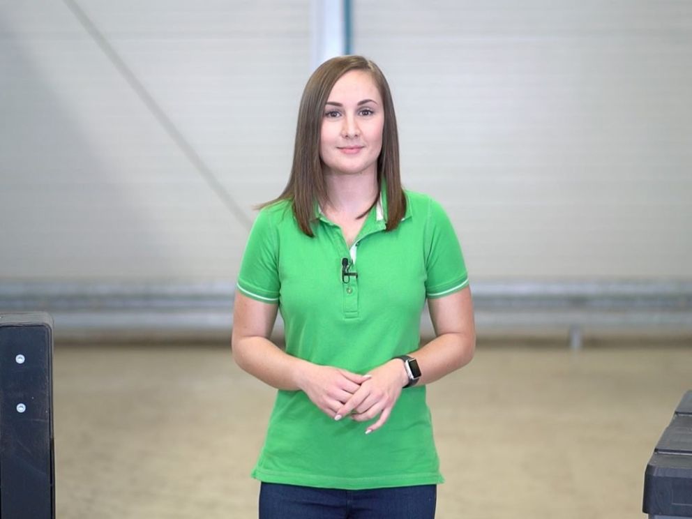 A person with shoulder-length hair, wearing a green polo shirt, stands confidently in a well-lit industrial environment.