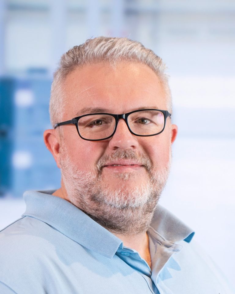 A man with glasses and gray hair, wearing a light blue polo shirt, smiles for the camera against a blurred background.