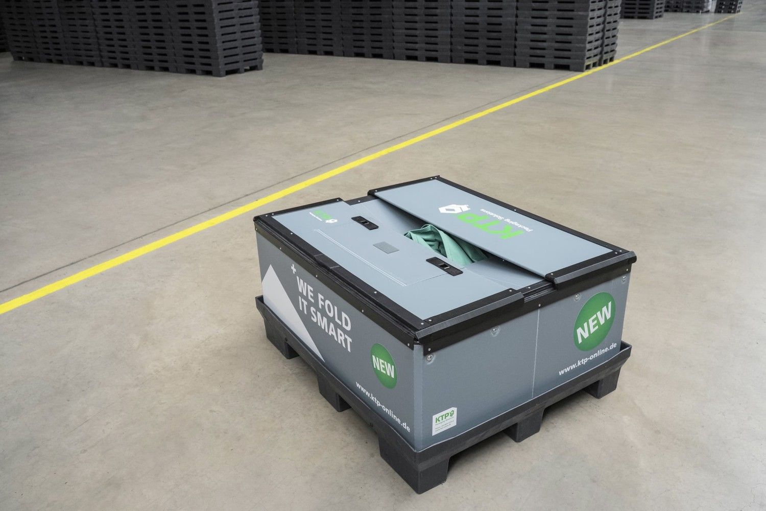 A foldable storage box with a green lid, labeled "WE FOLD IT SMART" and "NEW," placed on a concrete floor with a yellow line in a warehouse setting.