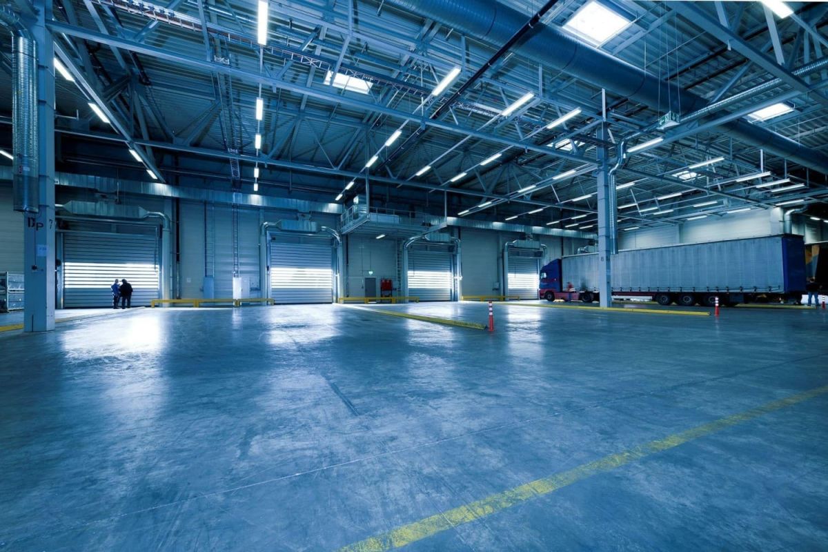 A spacious industrial warehouse interior with large loading doors, illuminated by overhead lights, featuring a parked truck and two people standing near the doors.