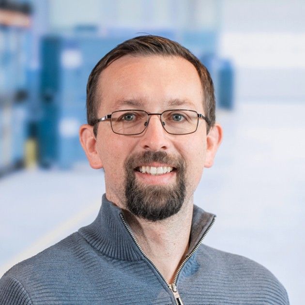 A smiling man with glasses and a beard, wearing a gray sweater, posing in a bright, blurred background.