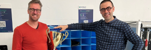 Two smiling men stand together, one holding a trophy, in front of a blue shelving unit with various compartments.