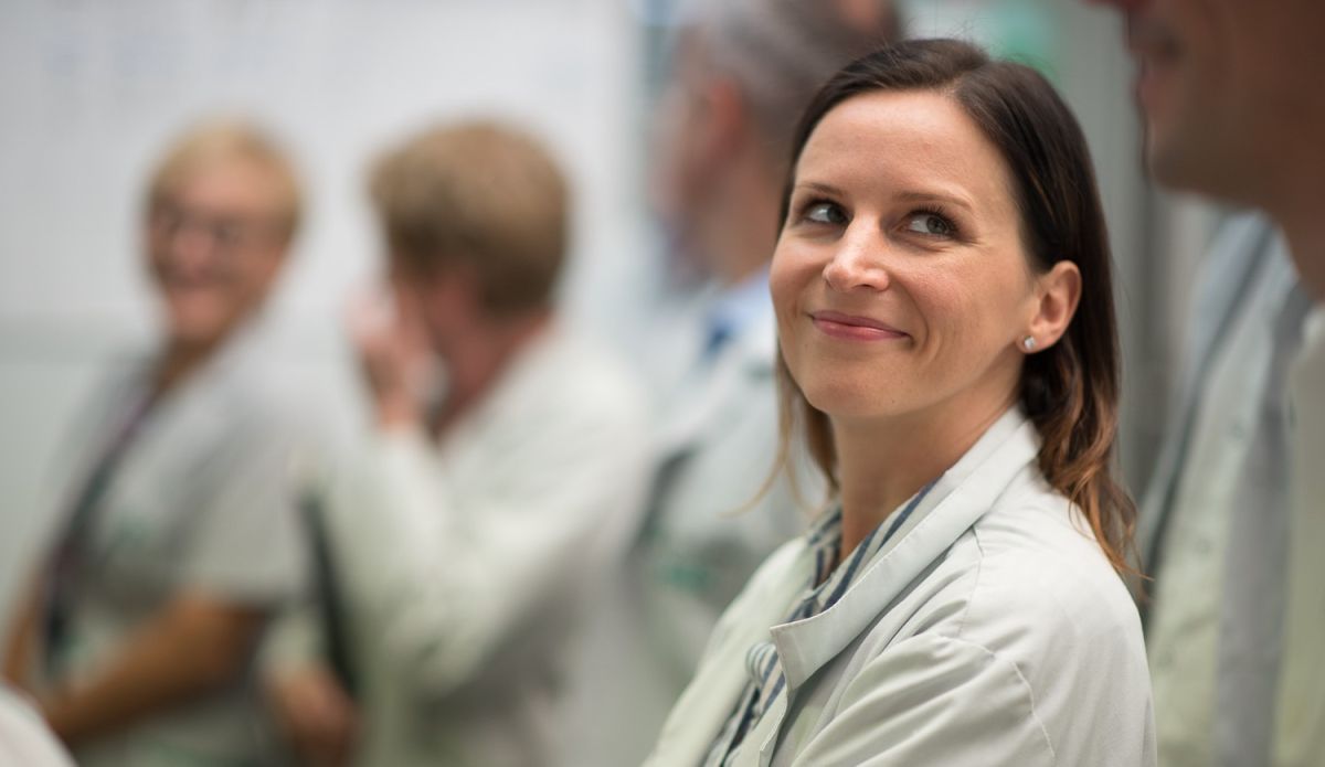 A female medical professional smiling while engaging in conversation with colleagues in a hospital setting.