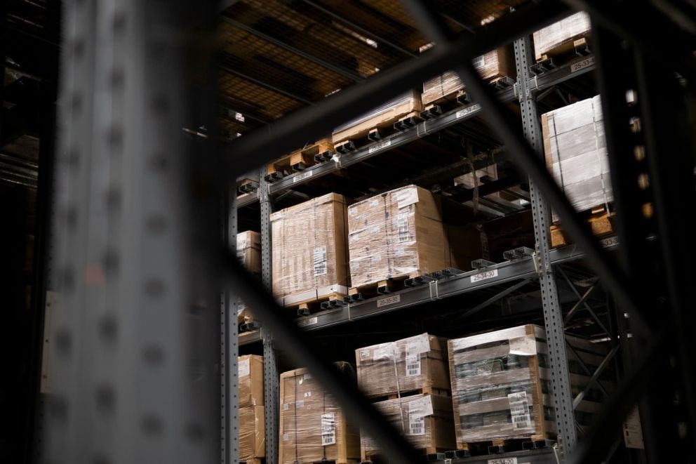 A warehouse storage area with stacked wooden pallets wrapped in plastic film on metal shelving, partially obscured by metal beams.