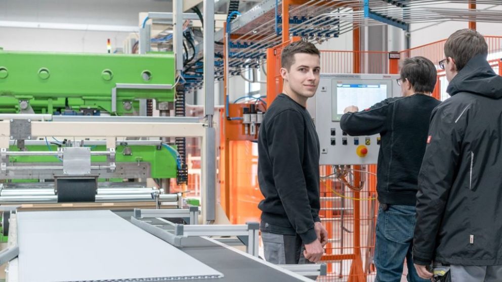 Three individuals are working in a factory environment, with machinery in the background. One person is facing the camera, while the other two are operating a control panel. The machinery features green and orange components, and there is a conveyor-like structure in the foreground.