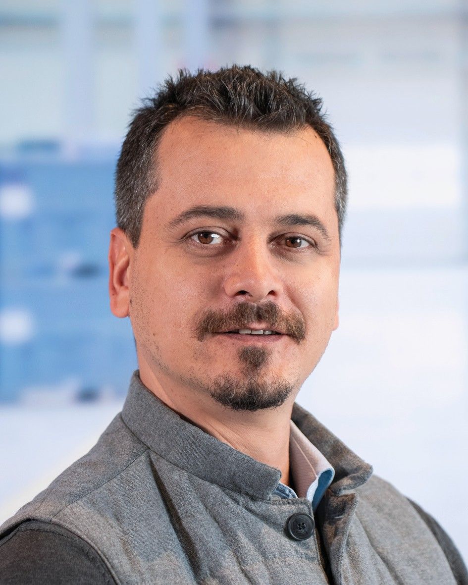 A man with short dark hair and a mustache, wearing a gray button-up vest, smiling softly against a blurred background.