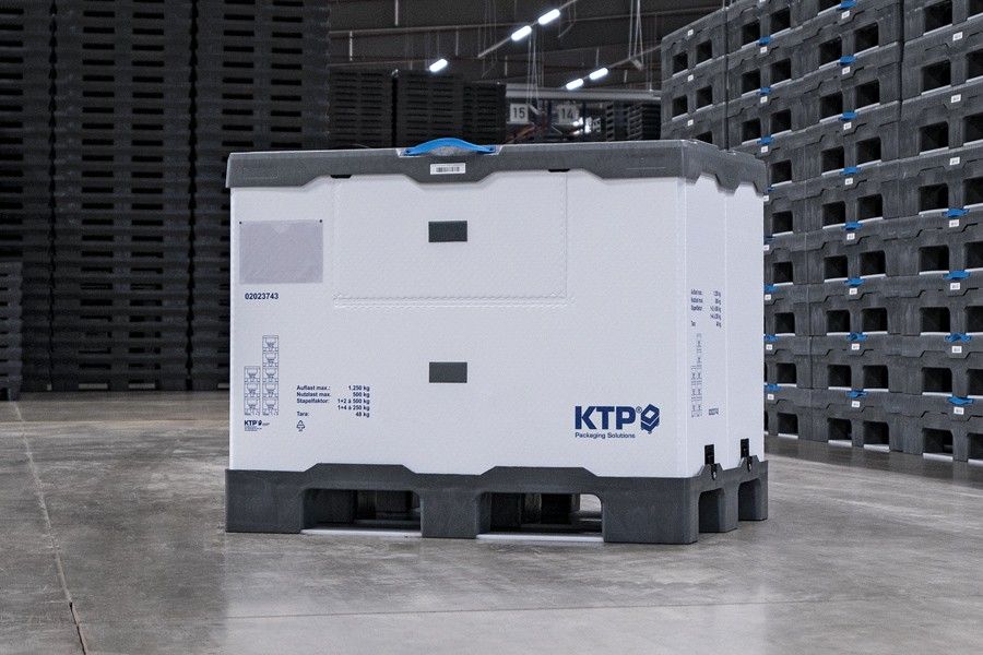 A large white packaging container with a gray base, labeled "KTP Packaging Solutions," situated in a warehouse among stacks of dark gray plastic pallets.