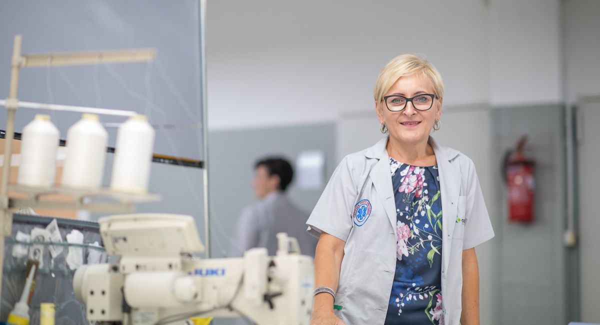 A smiling woman in a professional outfit stands beside a sewing machine in a workshop, with spools of thread and another person blurred in the background.