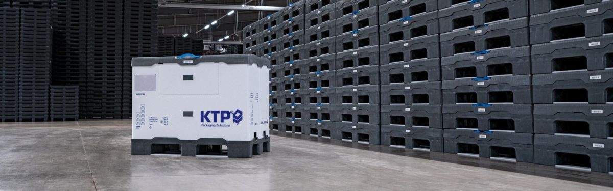 A large white and gray plastic container with the logo "KTP Packaging Solutions" is positioned in a warehouse aisle surrounded by stacked black plastic storage bins.