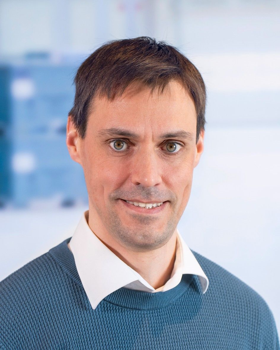 Portrait of a man with short dark hair, wearing a light blue sweater and a white collared shirt, smiling against a softly blurred background.