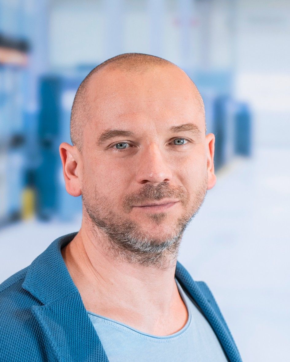 A man with a shaved head and stubble, wearing a blue blazer over a light shirt, posing against a blurred background of a modern indoor space.