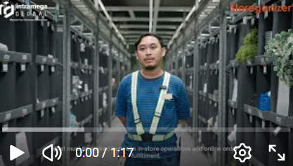 A man wearing a blue striped shirt and suspenders stands in a warehouse aisle lined with storage containers and plants.