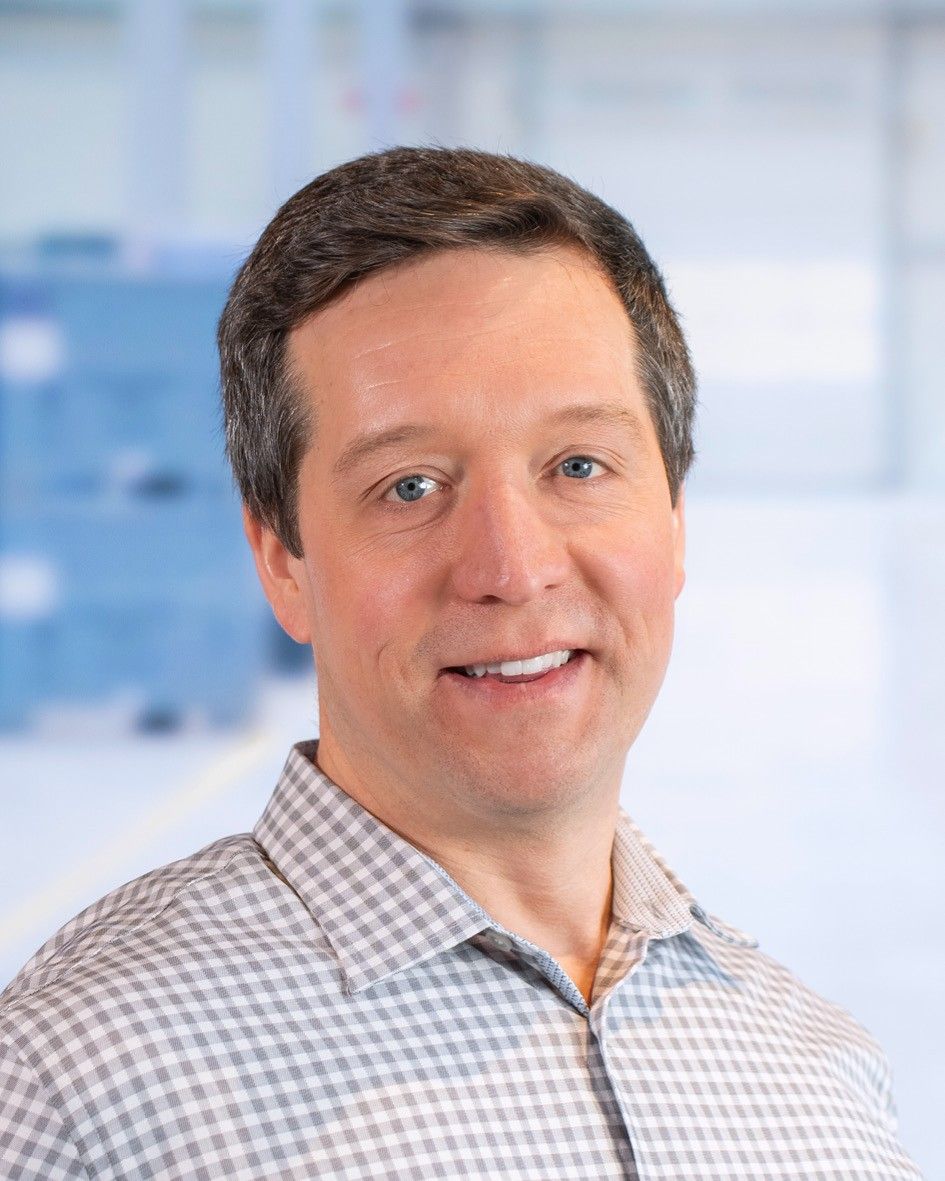 A smiling man with short hair wearing a checkered shirt, standing in an indoor setting with a blurred background.