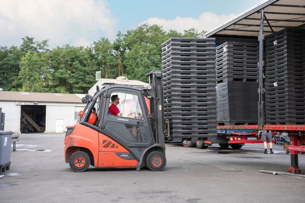 A forklift is moving near a truck loaded with stacks of black pallets in a warehouse environment, with trees and a building in the background.