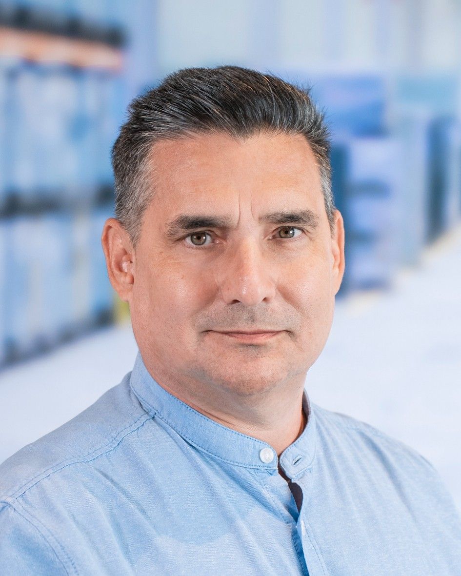 A portrait of a middle-aged man with short dark hair, wearing a light blue button-up shirt, against a blurred background of shelves.