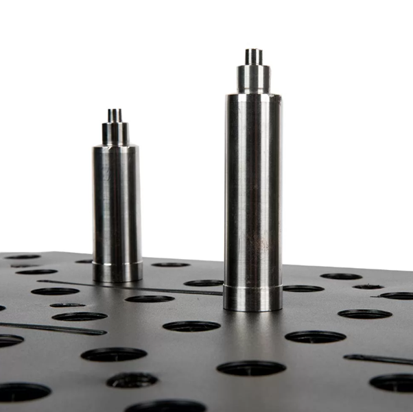 Two metal cylindrical pieces standing on a perforated black surface.
