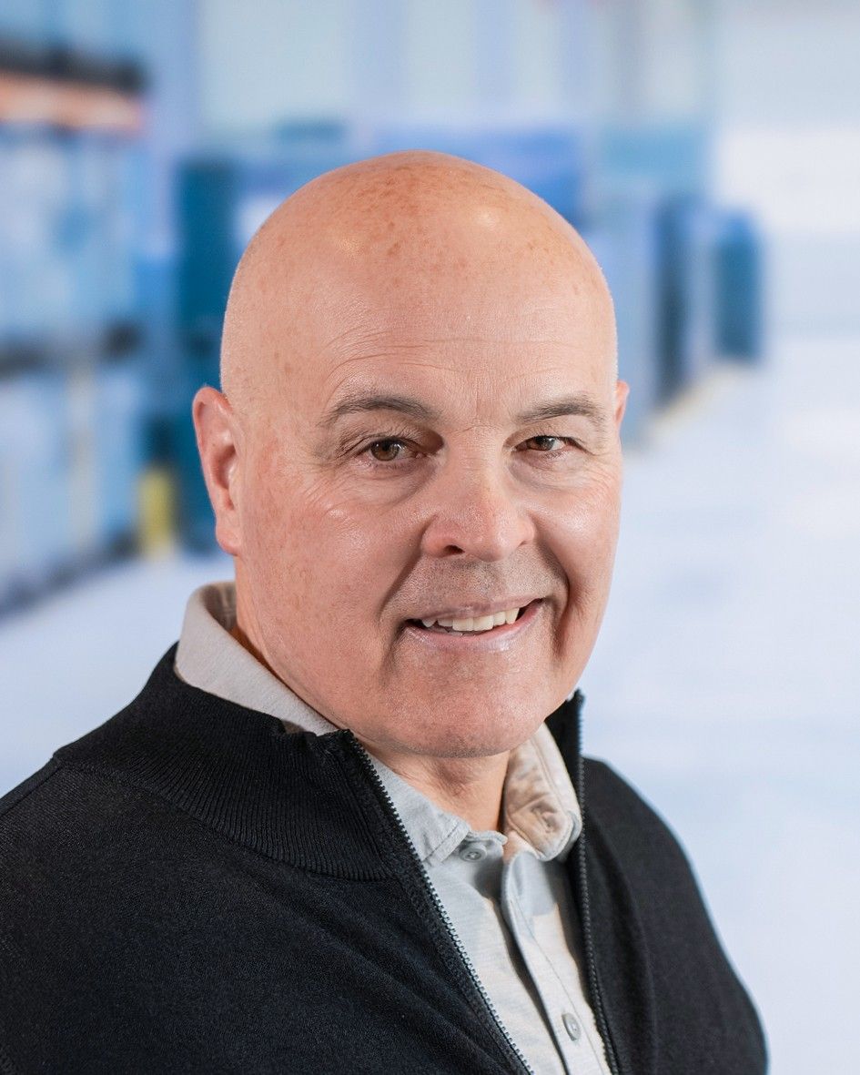 A smiling middle-aged man with a bald head, wearing a black zip-up sweater over a light grey collared shirt, with a blurred background of a warehouse setting.