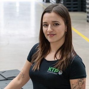 A woman with long brown hair wearing a black shirt with "KTP" printed on it, posing confidently in an industrial setting with pallets in the background.