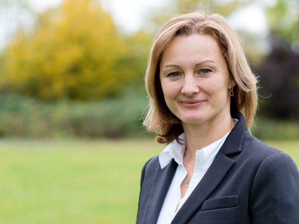 A confident woman in a business suit stands smiling outdoors with green grass and trees in the background.