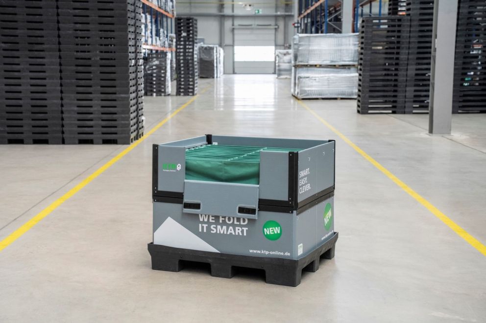 A foldable storage container with a green cover, labeled with the text "WE FOLD IT SMART," is placed in a well-lit warehouse aisle, surrounded by stacks of black pallets.