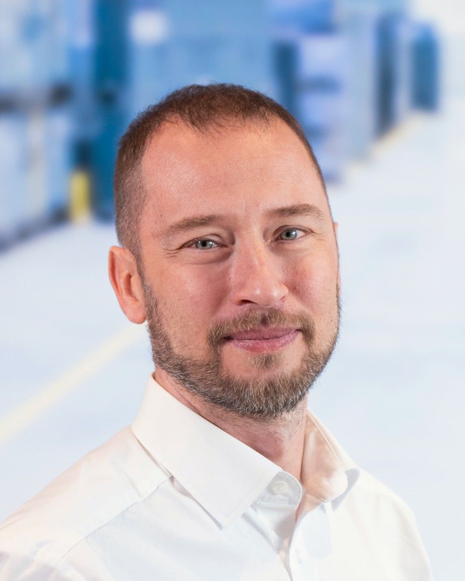 A man with a short beard and light-colored shirt smiling in a professional setting.