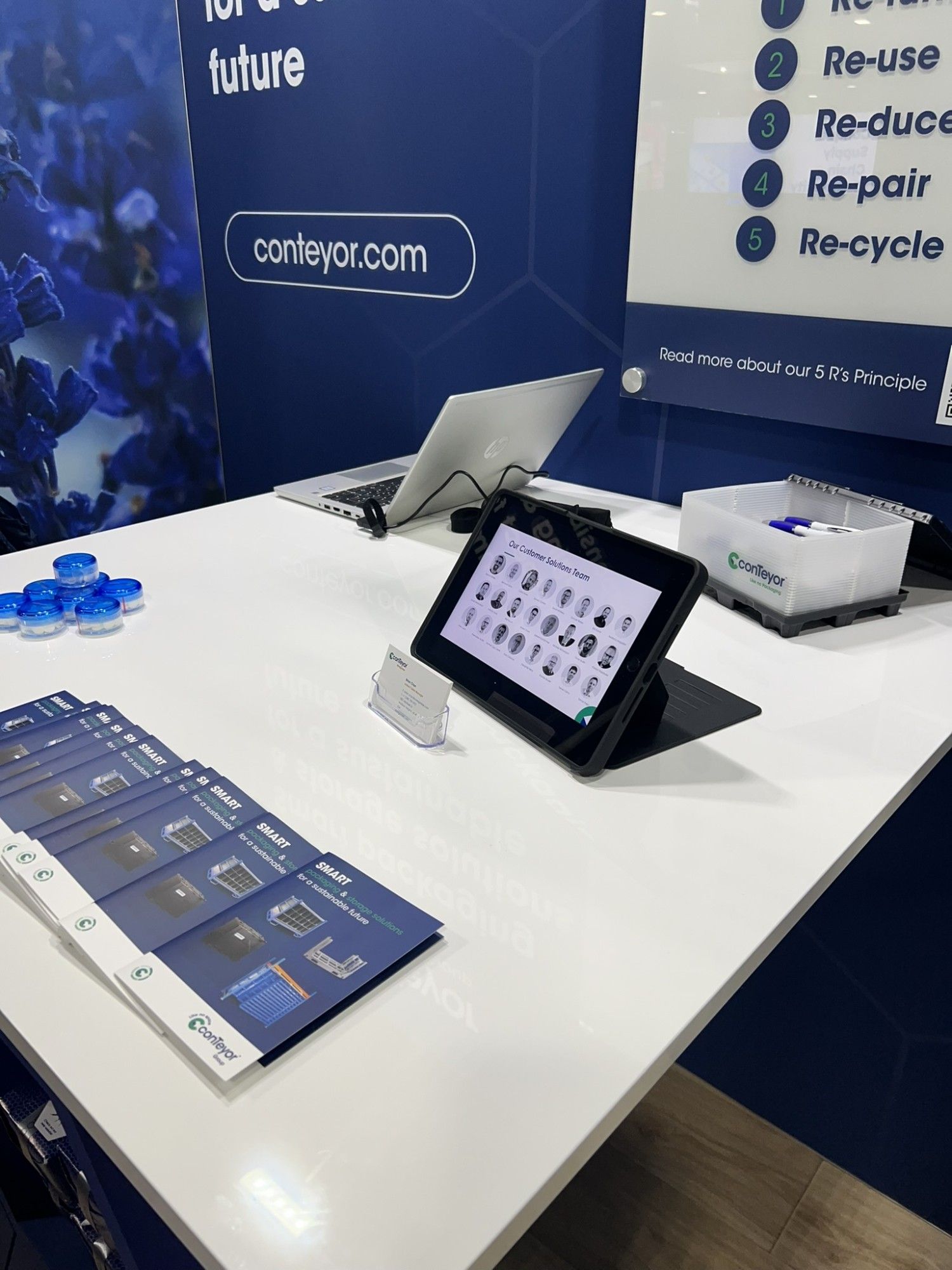 A trade show booth with a white table displaying brochures, a tablet showcasing images of products, a laptop, and promotional materials, set against a blue background featuring a company logo and sustainability principles.