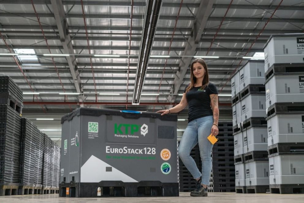 A woman in a black shirt stands next to a large gray packaging container labeled "EuroStack128" in a warehouse filled with stacked containers.