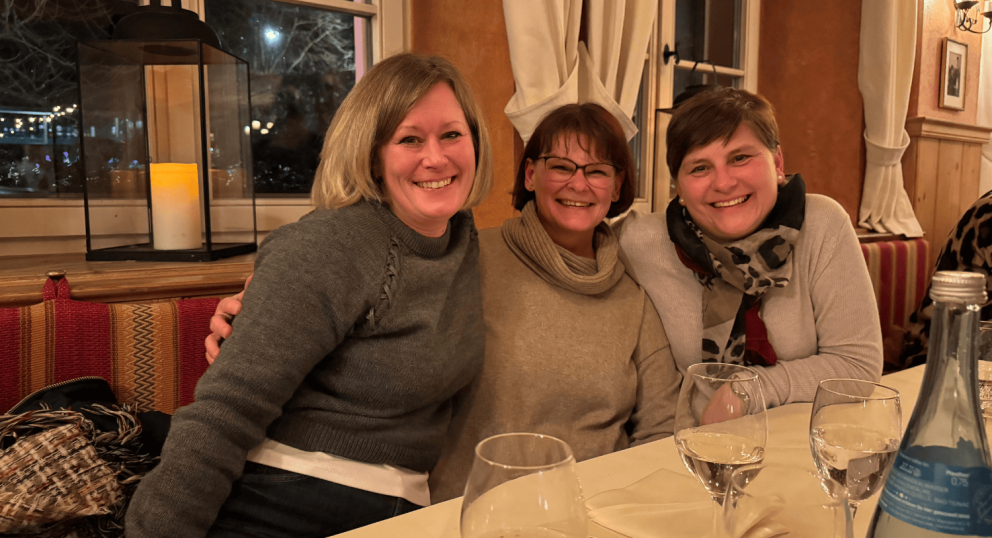 Three women smiling and posing together at a table with wine glasses, in a warm and cozy restaurant setting. A lit lantern is visible in the background.