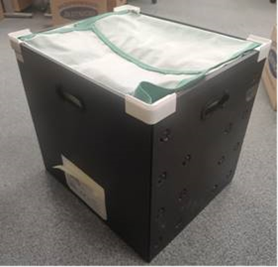 A black storage container with a green and white mesh cover, sitting on a gray carpeted floor, surrounded by boxes in a storage or office setting.