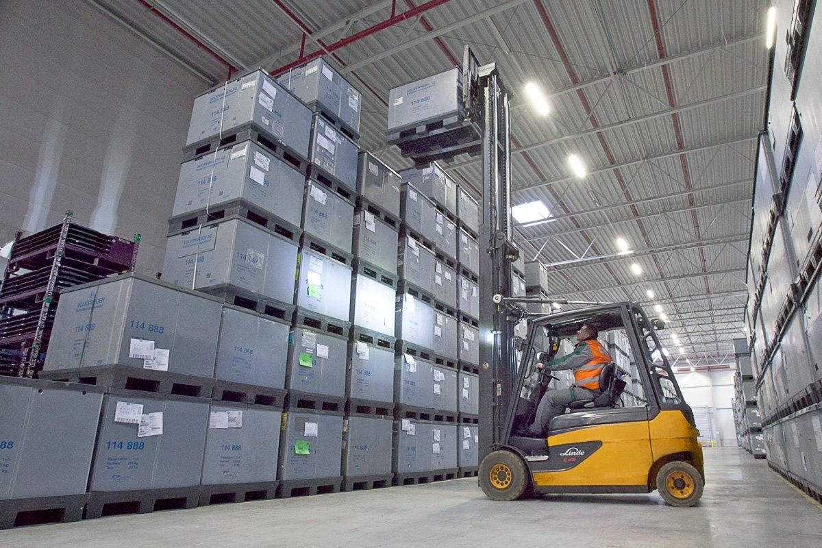 A forklift operator is maneuvering a lift to place a large box on top of a stacked palette in a warehouse.