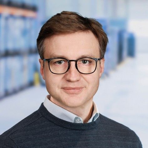 A man with glasses and short brown hair, wearing a dark sweater, smiles in a bright, blurred indoor setting.
