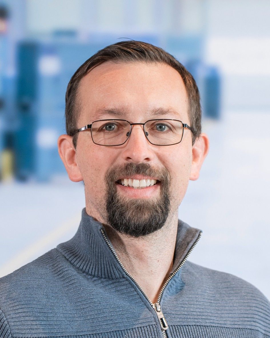 Smiling man with glasses and a beard, wearing a gray zip-up sweater, against a blurred background.