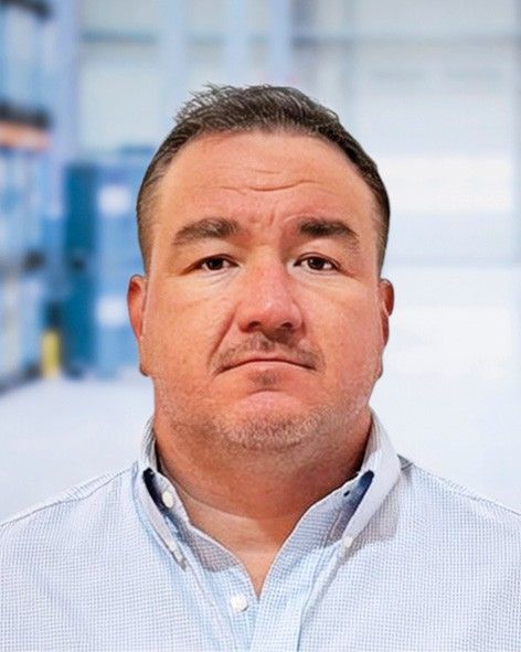 A man with short hair, wearing a light blue button-up shirt, stands in front of a blurred industrial background.