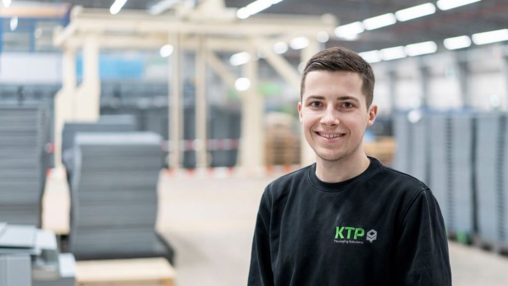 A young man smiling in a warehouse environment, wearing a black sweatshirt with "KTP" logo, surrounded by stacks of materials.