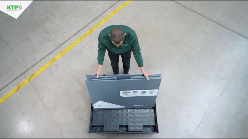 A person in a green jacket is demonstrating the setup of a packaging solution, standing over a black container with folded panels.