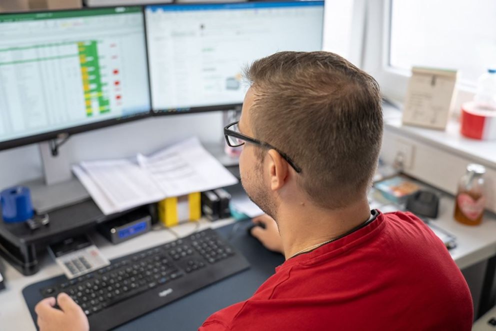 A man sitting at a desk working on a computer with dual monitors, showing spreadsheets and data.