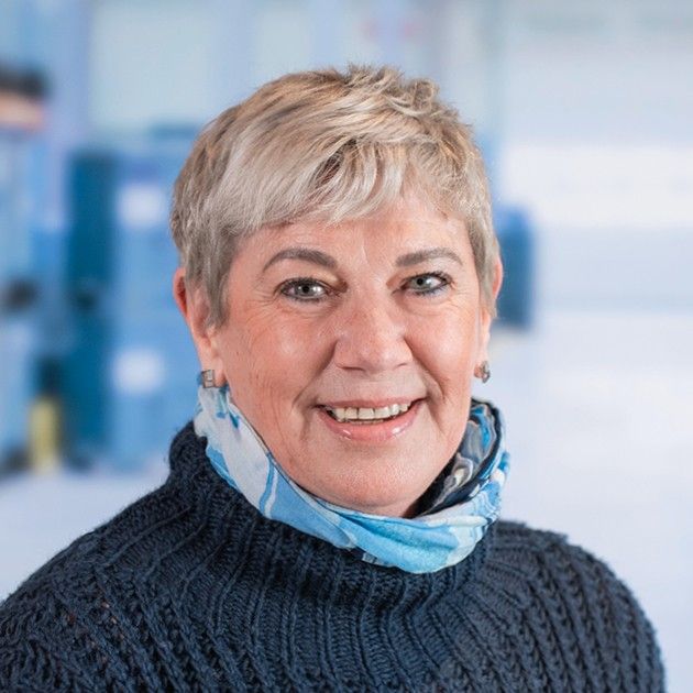 A smiling woman with short light-colored hair wearing a blue scarf over a dark sweater, posed against a blurred background.