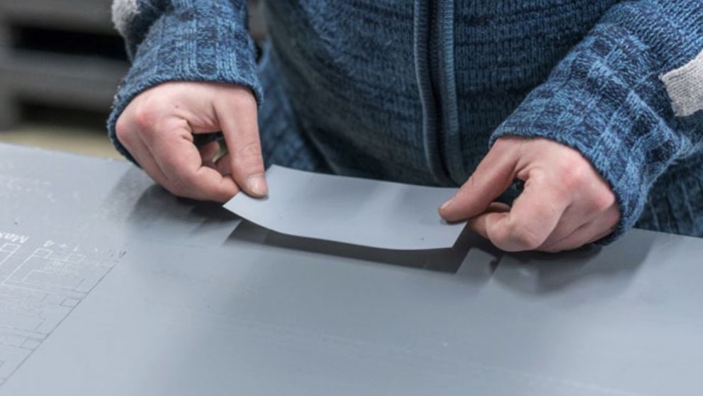 A person wearing a blue sweater is placing a gray sheet on a smooth surface.