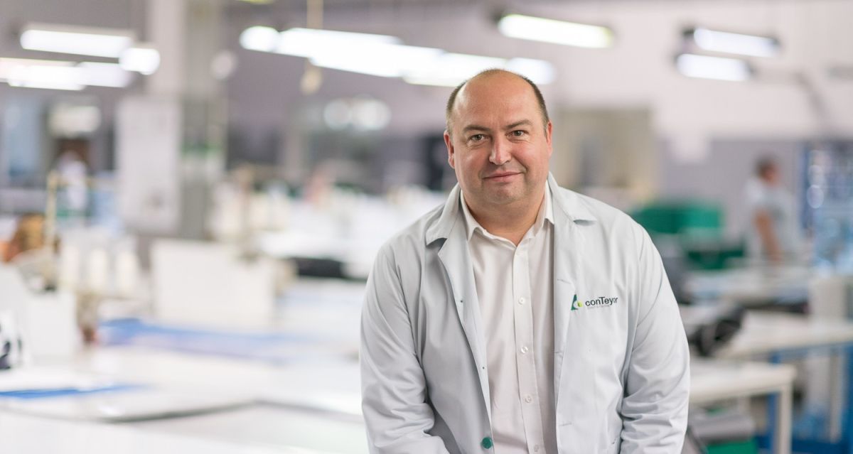 A man in a white lab coat stands in a bright industrial workspace with blurred workstations in the background.