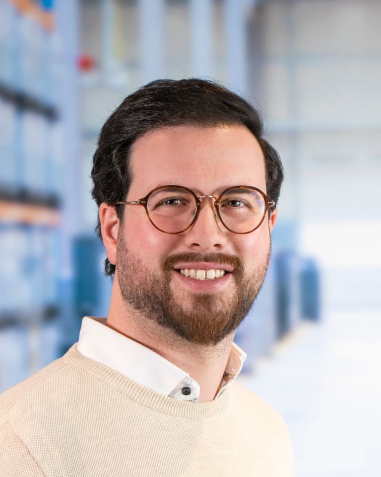 Smiling man with glasses and a beard, wearing a light sweater, standing against a blurred background of shelves or storage.