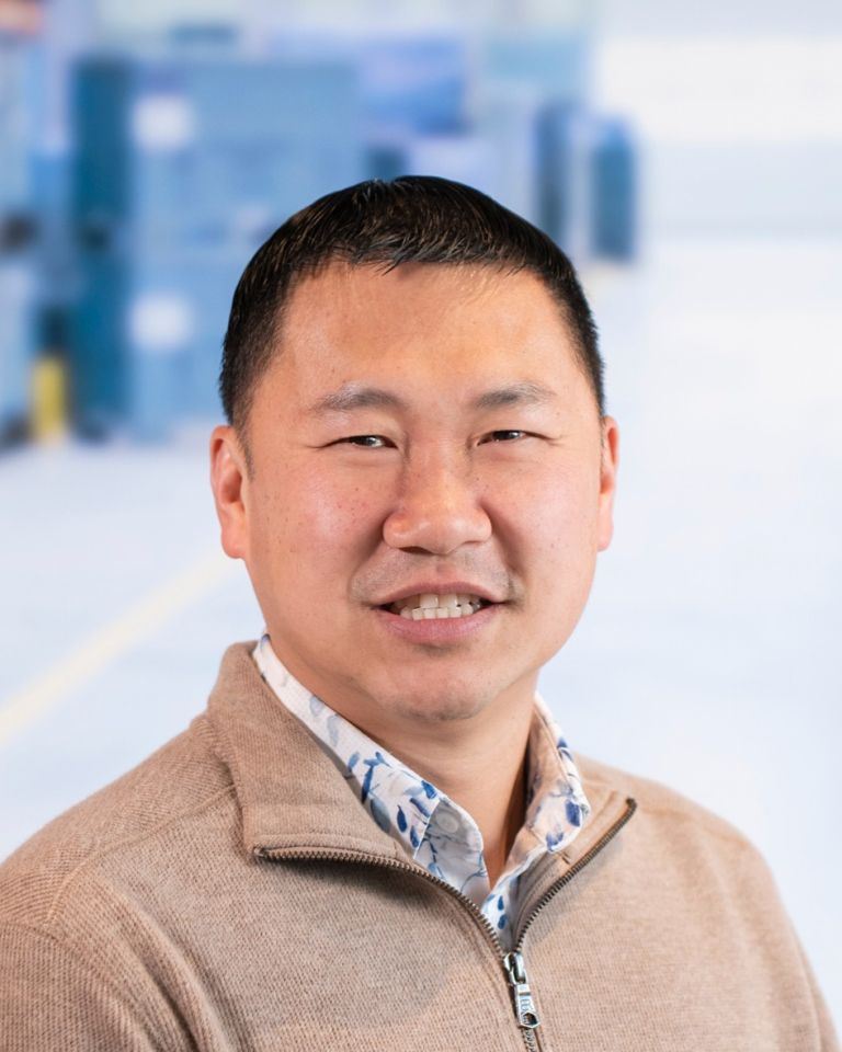 A close-up portrait of a man smiling, wearing a tan sweater and a patterned shirt, with a blurred background.