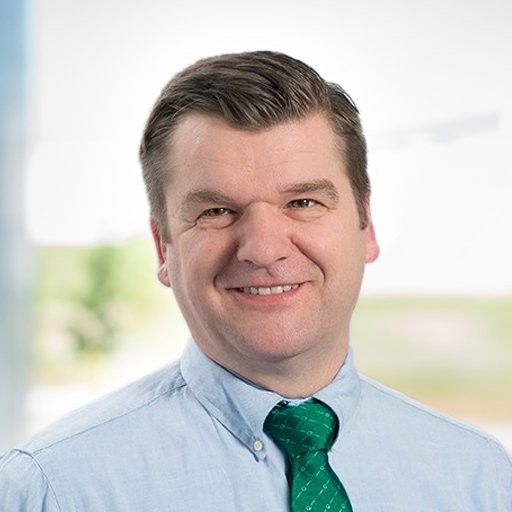 A smiling man in a light blue shirt and green tie, with a blurred outdoor background.