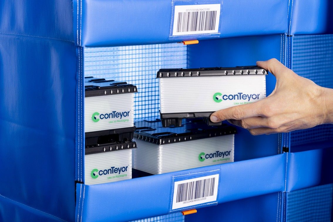 A hand reaching to place a conTeyor container onto a blue storage rack with barcode labels.