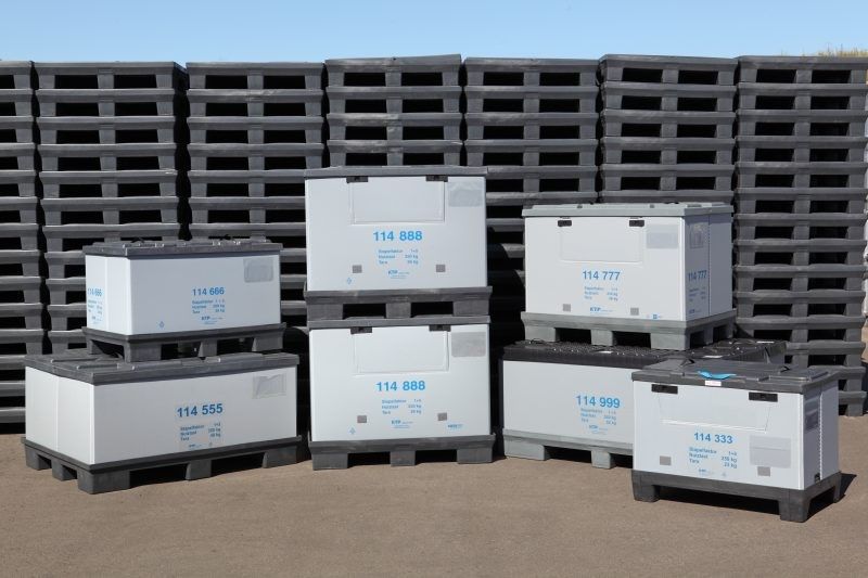 A collection of stacked shipping containers with gray bases and white tops, labeled with various numbers, set against a backdrop of more stacked pallets under a clear blue sky.