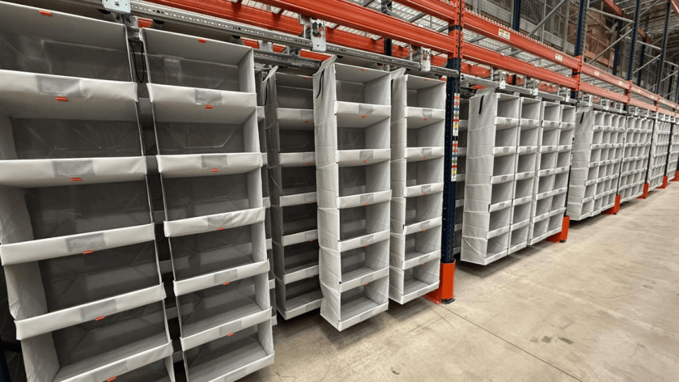 A row of empty storage bins on shelves in a warehouse setting, with a concrete floor and industrial shelving.